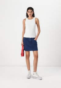 Biely rebrovaný top bez rukávov, modrá denimová sukňa a červená obdĺžniková kabelka. Modelka má na sebe biele vysoké tenisky, čím prezentuje ležérny outfit.