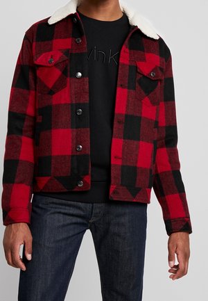 Mand iført rød og sortternet jakke med hvid fleecekrave over sort skjorte og mørkeblå jeans mod grå baggrund.