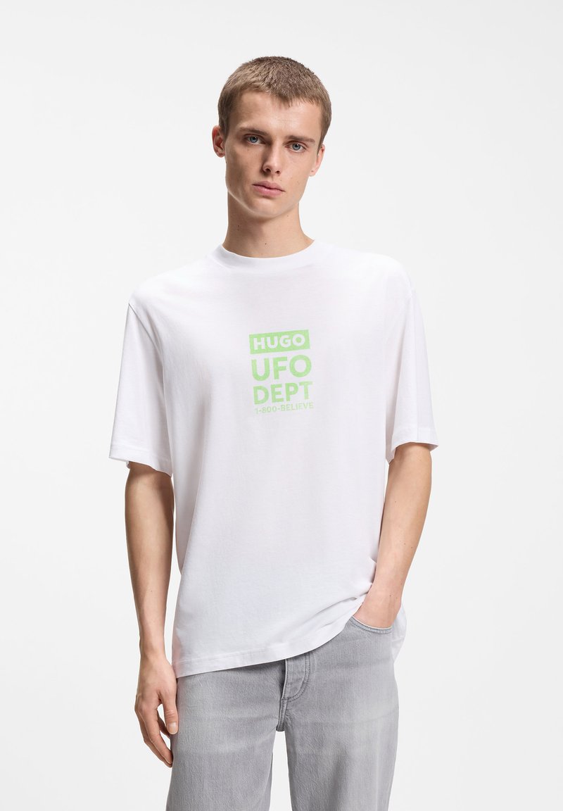 Νεαρός άνδρας στέκεται με το ένα χέρι στην τσέπη, φορώντας λευκό μπλουζάκι με πράσινο κείμενο "HUGO UFO DEPT 1-800-BELIEVE" και ανοιχτό γκρι τζιν.