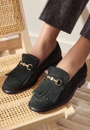 Dark green suede loafers with fringe detail, gold hardware accents, rounded toe, and a low heel, positioned on a woven chair.