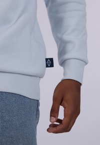 Felpa azzurra con polsini e orlo a costine. Presenta un piccolo etichetta blu navy con un logo geometrico sul lato. Jeans in denim visibili sotto.
