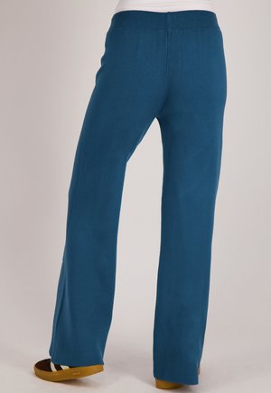 Persoon draagt blauwe wijde broek en bruine schoenen, staand op een effen lichte achtergrond.