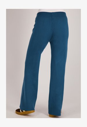 Persoon draagt blauwe wijde broek en bruine schoenen, staand op een effen lichte achtergrond.