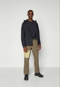Schwarze wasserdichte Jacke über einem dunklen Hemd, kombiniert mit beigen und khakifarbenen Hosen mit Seitentaschen. Der Moderator trägt Sonnenbrillen und schwarze Schuhe.