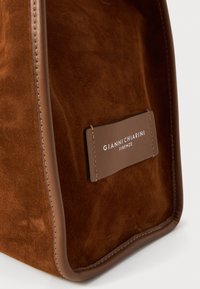 Sac à main en daim marron avec des accents en cuir lisse marron, présentant une étiquette avec du texte, une texture douce et un design minimaliste.