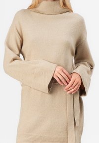 Pull col roulé beige, texture en maille douce, coupe ample, manches larges, détails côtelés aux poignets et à l'ourlet, ceinture en tissu à nouer à la taille.