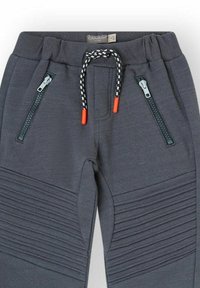 Los pantalones de chándal grises presentan un diseño texturizado con rayas diagonales, dos bolsillos con cremallera, una cinturilla elástica y un cordón con puntas naranjas.