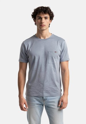 Giovane uomo con capelli ricci che indossa una t-shirt azzurro chiaro con tasca e jeans azzurri chiari, in piedi davanti a uno sfondo semplice.