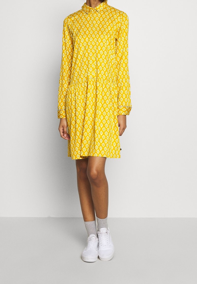 Personne portant une robe jaune à manches longues avec des motifs losanges blancs, des chaussettes grises aux chevilles et des baskets blanches, debout sur un fond uni.
