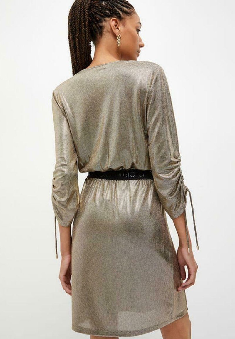 Gouden, metallic jurk met lange mouwen, trekkoorddetails aan de zijkanten en een zwarte riem die de taille accentueert. Soepele textuur.