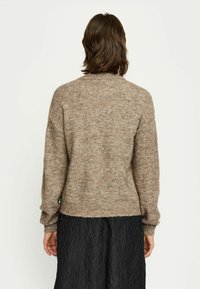 Pull en maille marron avec une coupe décontractée, un col rond et des poignets côtelés. La texture semble douce et légèrement chinée. Porté sur une jupe noire texturée.