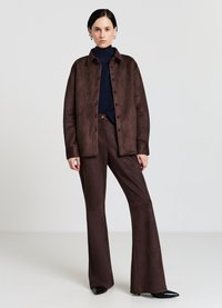 Braunes Oberteil aus Wildleder mit Knöpfen, kombiniert mit passenden ausgestellten Hosen. Darunter ein blaues, geripptes Rollkragenpullover. Schwarze spitze Schuhe. Glatte Textur.