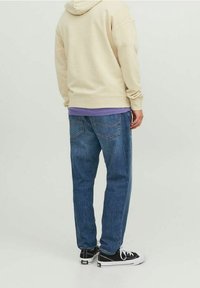 Beige huvtröja över en lila skjorta, kombinerad med blå denimjeans och svarta sneakers. Synliga sömmar i bakfickan.