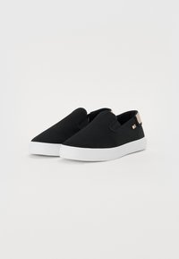 Schwarze slip-on skor med ovandel i kanvas, vit gummisula och en liten beige accent vid hälen. Minimalistisk design, platt form.