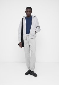 Sweat à capuche gris zippé sur un t-shirt bleu, assorti à un pantalon de survêtement gris et des chaussures noires. Un sac noir texturé est porté en bandoulière.