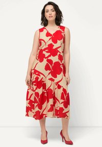 Beige květinové šaty s červenými přehnanými květinami, s výstřihem do V, bez rukávů a s vrstveným volánkovým lemem. Doplňují je červené vysoké podpatky.