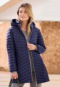 Marineblauwe geïsoleerde jas met een doorgestikt patroon, capuchon, ritssluiting en witte accentpiping. Draag het over een grijze top.