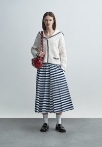Donna che indossa un cardigan bianco testurizzato con finiture blu navy, una gonna midi a righe blu e bianche, calzini bianchi, mocassini neri e che porta una borsa rossa.