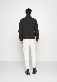 Black quilted jacket with a high collar, featuring shoulder seams and embroidered logo on the back. Model wears white pants and black boots.