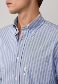Hombre vestido con camisa de botones a rayas verticales en azul claro y blanco, con un bolsillo frontal y botones en el cuello, mirando hacia la izquierda.