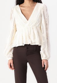 Blusa bianca in pizzo con scollatura a V profonda e maniche lunghe a palloncino gatherate; materiale trasparente con ricami floreali; abbinata a pantaloni marroni.