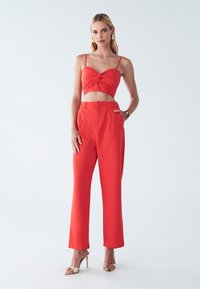 Červený dvojdielny outfit pozostávajúci z vykrojeného, skrúteného topu s tenkými ramienkami a širokými, prispôsobenými nohavicami. Hladký materiál; minimalistický dizajn.