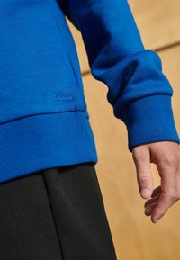 Felpa blu a maniche lunghe con polsini a costine, dotata di un sottile logo Clarks. Indossata sopra un tessuto nero con una superficie liscia.