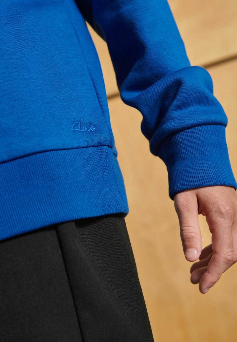 Felpa blu a maniche lunghe con polsini a costine, dotata di un sottile logo Clarks. Indossata sopra un tessuto nero con una superficie liscia.