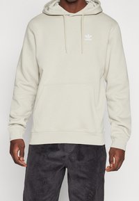 Muž stojící proti jednoduchému pozadí v světle béžové mikině Adidas s přední kapsou a tkaničkami, v tmavých kordových kalhotách.