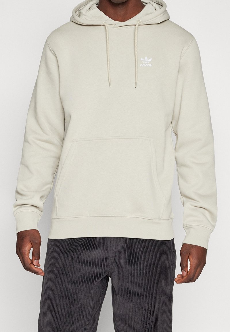 Muž stojící proti jednoduchému pozadí v světle béžové mikině Adidas s přední kapsou a tkaničkami, v tmavých kordových kalhotách.