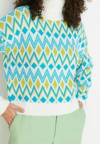 Vrouw in een witte coltrui met blauwe en groene diamant- en zigzagpatronen, gecombineerd met lichtgroene broek, met handen in de zakken.