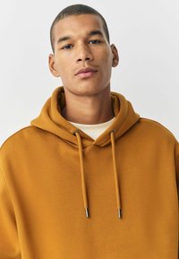 Sudadera color mostaza hecha de tela suave. Cuenta con una gran capucha, cordones con puntas de metal y un escote tipo crew sencillo.