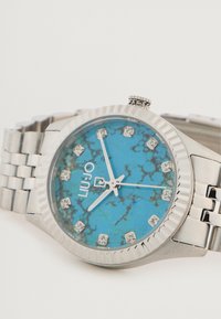 Sølv metalur ur med en turkis urskive, der har et marmorliknende mønster, krystal timemarkører og en struktureret bezel. Brandlogoet er vist.