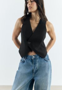 Schwarze ärmellose Wickelweste mit einem einzigen Knopfverschluss, kombiniert mit lockeren hellblauen Jeans, die einen modernen, strukturierten Look schaffen.