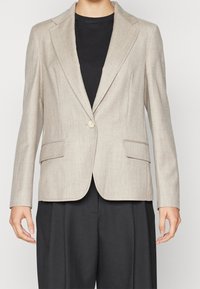 Beige blazer dengan finishing bertekstur, lapel terpotong, penutupan dengan satu kancing, dan dua kantong depan, dipakai di atas kemeja hitam.