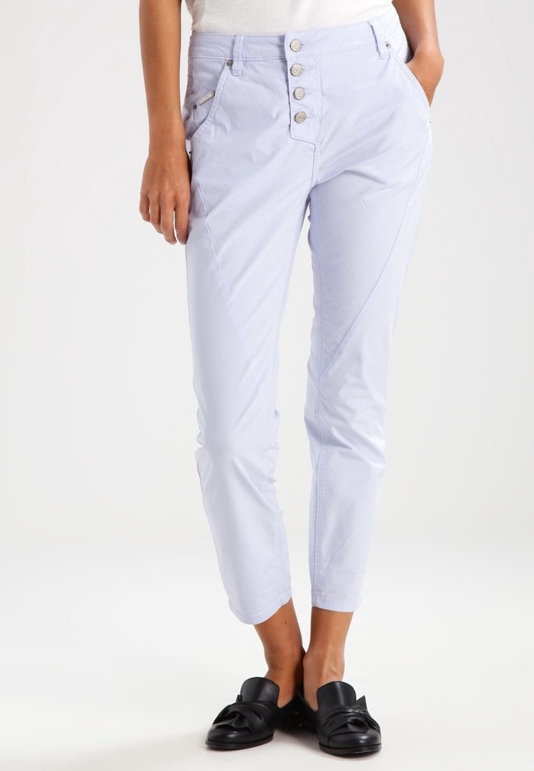 Femme portant un pantalon slim violet clair à quatre boutons sur le devant et des chaussures noires slip-on, debout les mains dans les poches.