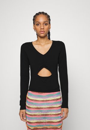 Μαύρη μπλούζα με μακριά μανίκια, V-λαιμό και κεντρικό άνοιγμα, συνδυασμένη με μία πολύχρωμη πλεκτή φούστα με ρίγες που διαθέτει ζωντανές υφές και σχέδια.