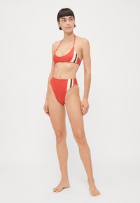 Sarkans bikini komplekts ar trīsstūra veida krūšturi un augsta jostasvieta apakšdaļu ar vertikālām brūnām un krēmkrāsas svītrām. Gluda auduma, minimāls aksesuārs.