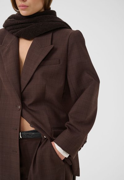 Femme en costume marron à carreaux avec blazer et pantalon, portant une écharpe noire texturée et une ceinture noire, main dans la poche.