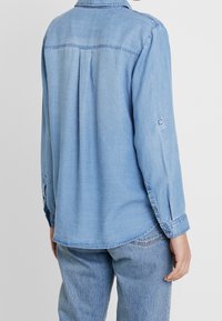 Light blue denim shirt, long sleeves, buttoned cuffs, and a curved hem. Features back pleat for added comfort and fit.