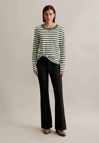 Mulher vestindo uma camisa de mangas longas verde e branca com riscas, calças pretas largas e sapatos castanhos, de pé contra um fundo bege liso.