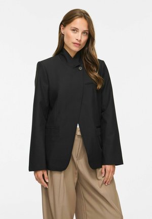Frau mit langen Haaren, die eine schwarze Knopfjacke und beige, weite Hosen vor einem schlichten hellen Hintergrund trägt.