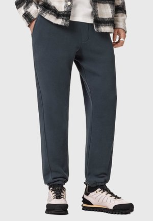 Person trägt dunkelblaue Jogginghose, weiße und schwarze Sneakers, weißes Hemd und schwarz-weiß karierte Jacke, Hände teilweise in den Taschen.