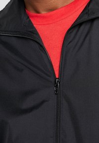 Svart jacka med dragkedja framtill, tillverkad av strukturerat material, bärs över en röd skjorta. Har krage och en figurnära design.