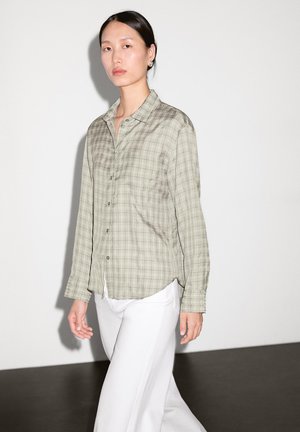 Light green plaid shirt with long sleeves, button-down front, and a curved hem, paired with white wide-leg pants against a neutral background.