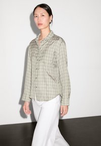 Light green plaid button-up shirt with long sleeves, rounded hem, and a relaxed fit, paired with white wide-leg trousers.