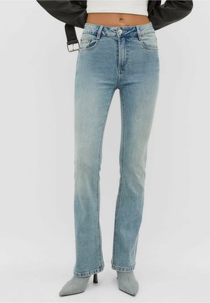 Kvinde iført lyseblå straight-leg jeans, grå spidse hæle, hvid cropped top og sort jakke med synlig bæltespænde.