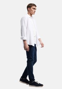 Valkoinen pitkähihainen paita valkoisen T-paidan päällä, yhdistettynä tummiin farkkuihin ja mustiin urheilukenkään. Ominaisuuksiin kuuluu käännetyt hihansuut ja rento istuvuus.