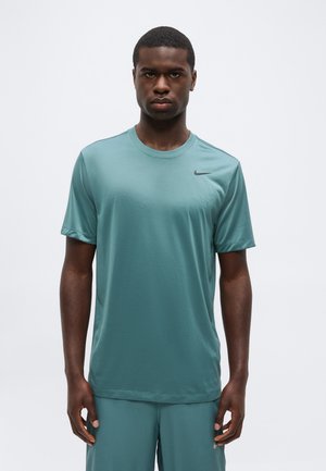 Mladenič v turkizni Nike športni majici in kratkih hlačah stoji pred enobarvnim belim ozadjem, obračajoč se naprej z nevtralnim izrazom.