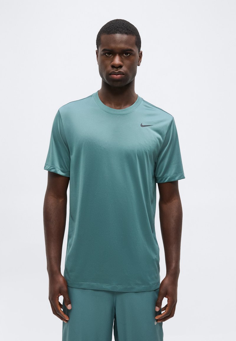 Νεαρός άνδρας φορώντας τιρκουάζ αθλητικό πουκάμισο και σορτς Nike, στέκεται μπροστά σε απλό λευκό φόντο, κοιτώντας προς τα εμπρός με ουδέτερη έκφραση.